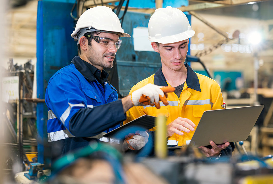 Two men in protective clothing with safety helmets, the older one explaining something to the younger one.