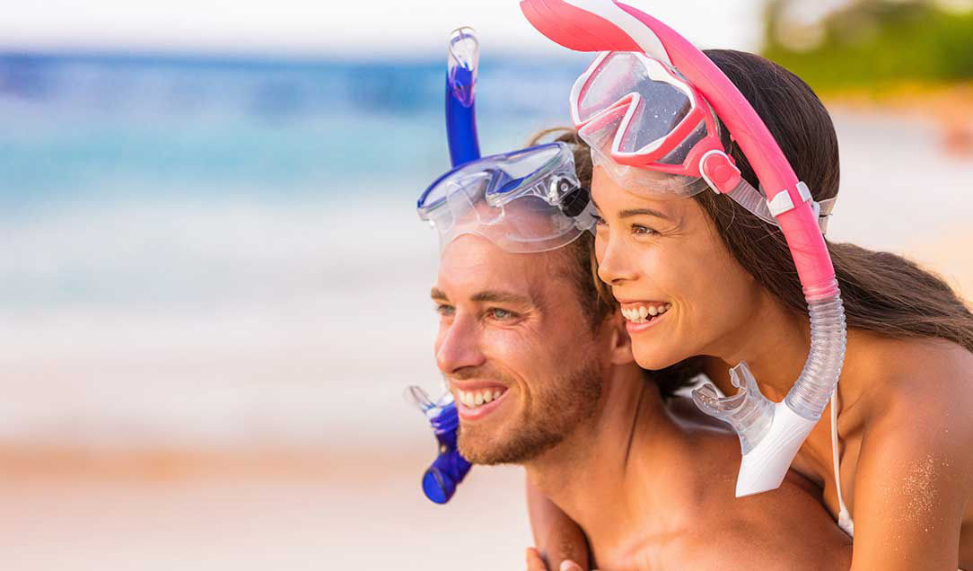 A man and woman with snorkels and goggles on the beach. They are smiling and looking into the distance.