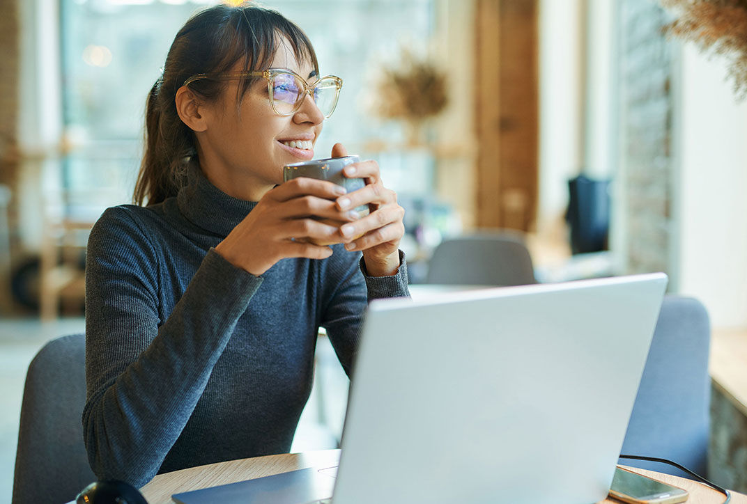 An employee is sitting at her work laptop and drinking from a cup.