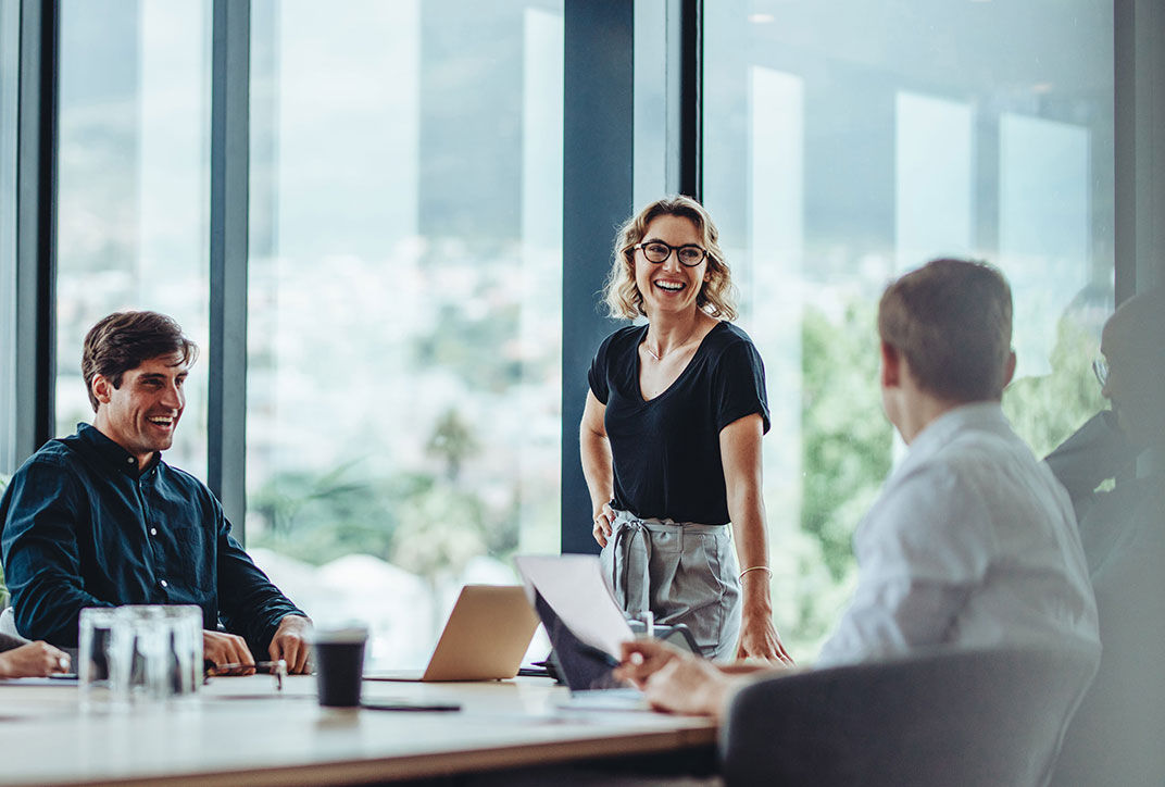 alt="A woman stands in a meeting in front of colleagues.