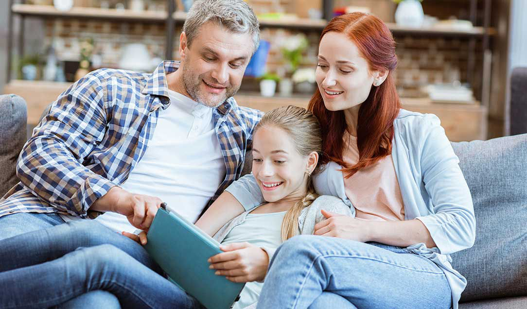 A man, a woman and a girl are sitting on a sofa, looking at something on a tablet.