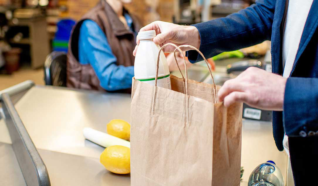 A supermarket checkout. In the background is the cashier; in the foreground a man is putting a bottle of milk into a paper