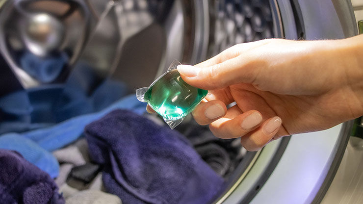 Laundry detergent pod held near an open washing machine drum filled with clothes